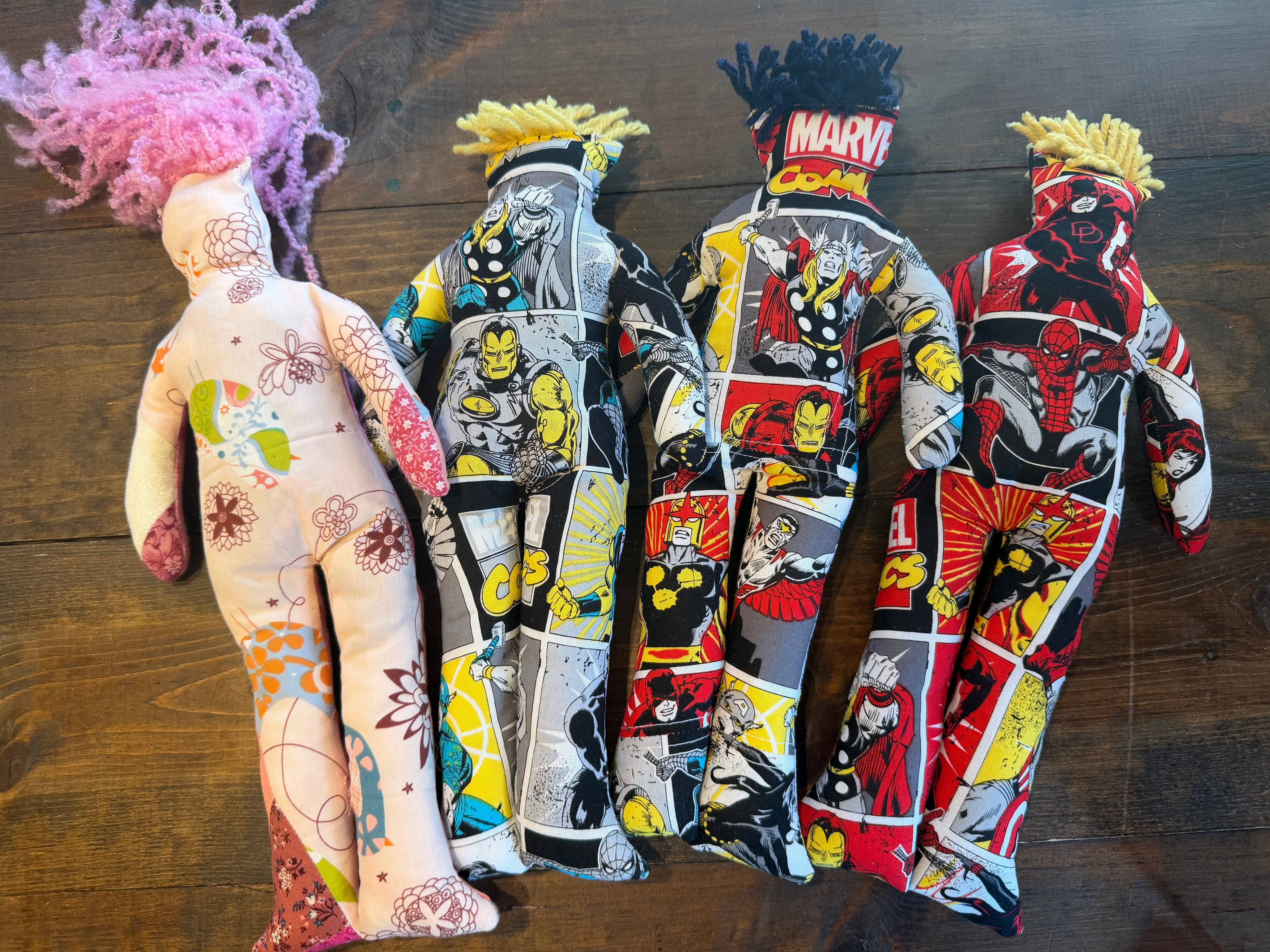 Three handmade cloth dolls with comic book character prints are arranged on a dark wooden floor. The doll at the top has a light pink body with multicolored floral patterns and pink yarn hair.