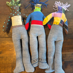Three handmade fabric puppets made from denim and patchwork fabrics, laid out on a dark wood floor. The puppets have gray denim legs and are decorated with colorful patchwork bodies in blue, yellow, pink, and brown.