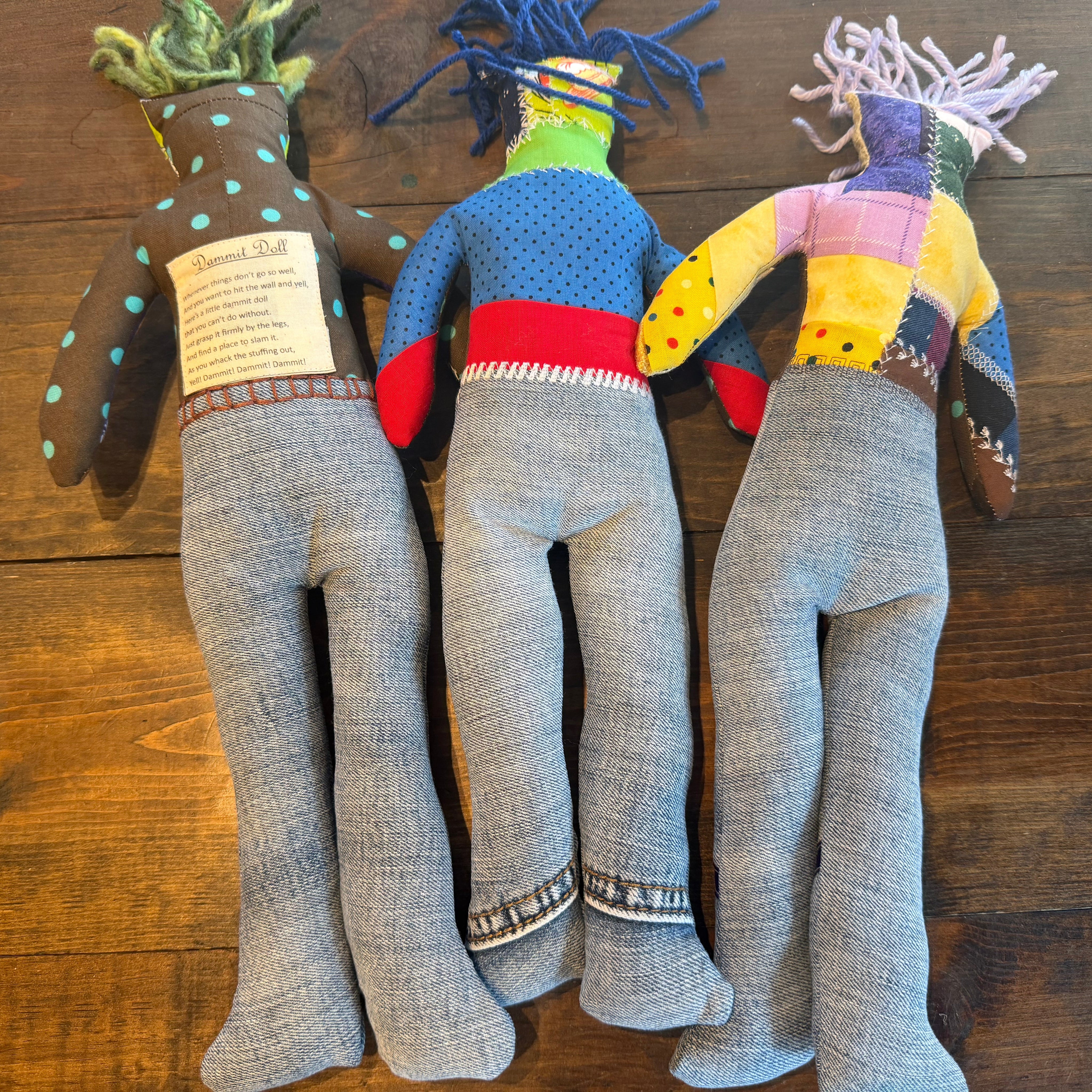 Three handmade fabric puppets made from denim and patchwork fabrics, laid out on a dark wood floor. The puppets have gray denim legs and are decorated with colorful patchwork bodies in blue, yellow, pink, and brown.